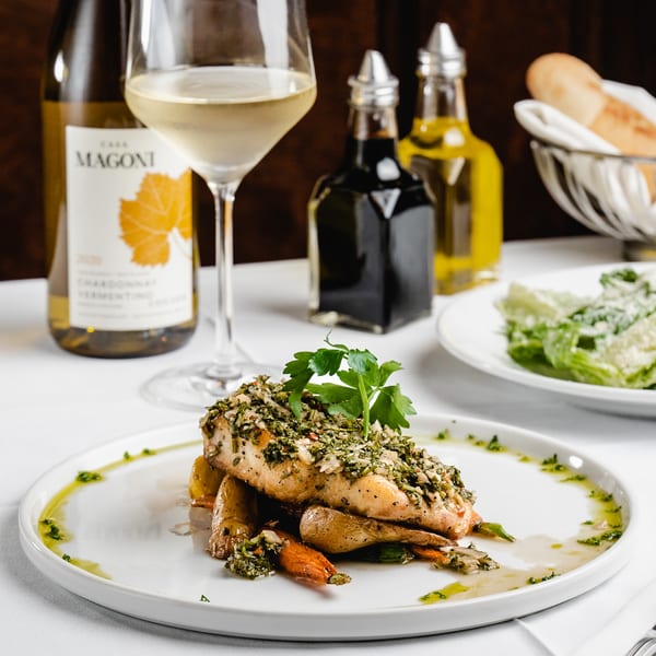 Herb-crusted fish with roasted vegetables, wine, and bread on a white tablecloth at Romesco Mexiterranean Bistro.