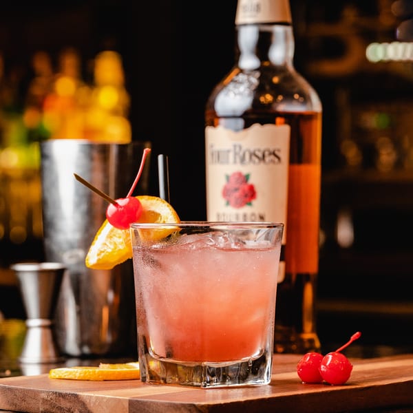A pink cocktail with orange and cherry garnish, next to a bourbon bottle and bar tools.