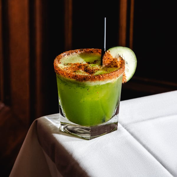 Green Romesco Mexiterranean cocktail with a chili-lime rim and cucumber slice on a white tablecloth.