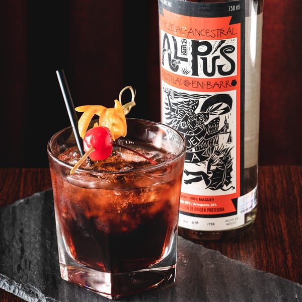 A dark cocktail with a cherry and orange peel garnish next to a bottle of Mezcal Ancestral.