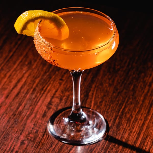 A Romesco Mexiterranean Bistro cocktail, garnished with a lemon slice and chili-salt rim, on a dark wooden table.