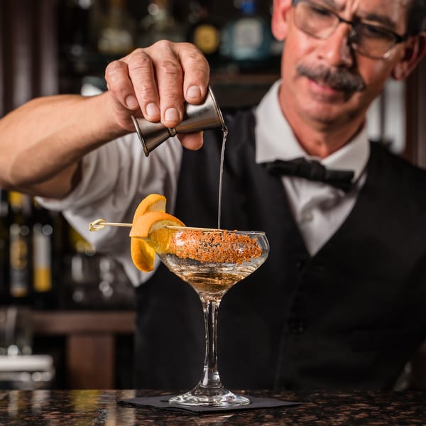 Bartender pouring a drink into a Romesco Mexiterranean Bistro cocktail glass with an orange peel garnish.