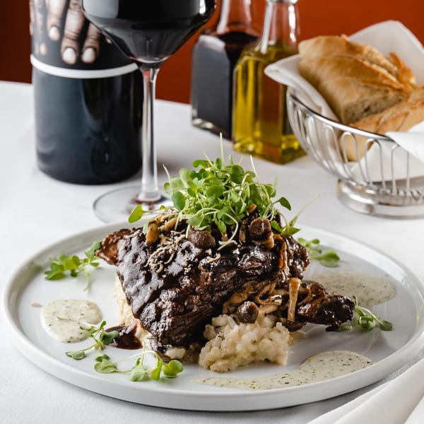 Braised short rib with mushrooms and microgreens on risotto, with wine and bread in the background.