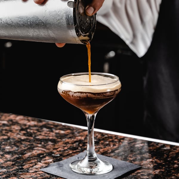Bartender pouring a dark, frothy cocktail from a shaker into a coupe glass on a granite bar.