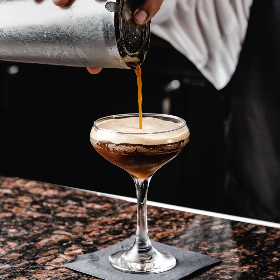 Bartender pouring a dark, frothy cocktail from a shaker into a coupe glass on a granite bar.