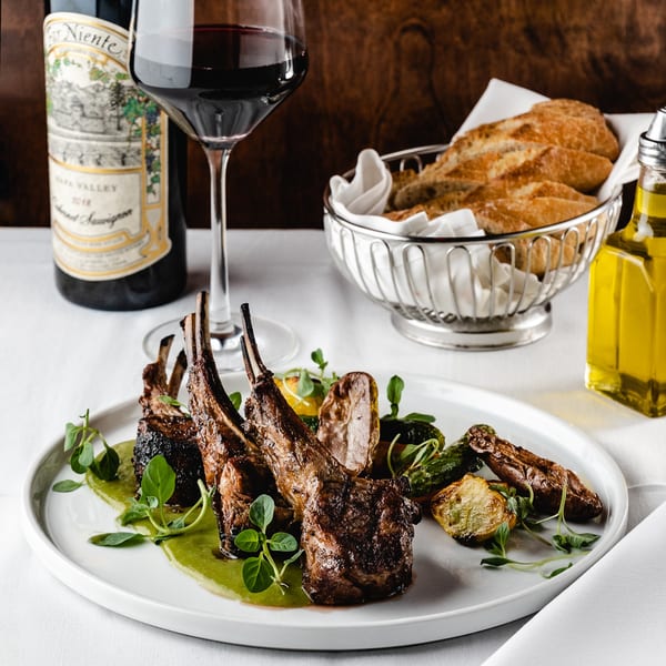 Grilled lamb chops with green sauce, roasted vegetables, wine, bread, and olive oil on a white tablecloth.