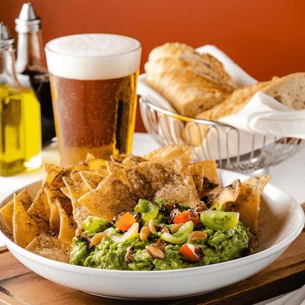Guacamole with tortilla chips, a glass of beer, and bread in a basket on a wooden table.