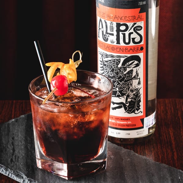 A dark cocktail with ice, an orange peel, and a cherry, next to a bottle of Mezcal Ancestral.