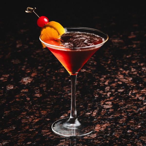 A dark red cocktail in a martini glass, garnished with an orange peel and a cherry.