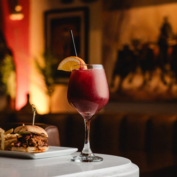 A vibrant red Romesco Mexiterranean Bistro drink with an orange slice and cherry, next to a slider.
