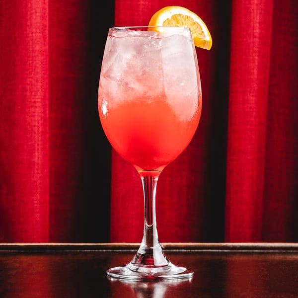 A vibrant red cocktail with ice and an orange slice, set against a red curtain background.