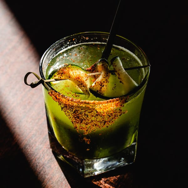 A vibrant green Romesco Mexiterranean Bistro cocktail, garnished with cucumber slices and chili seasoning.