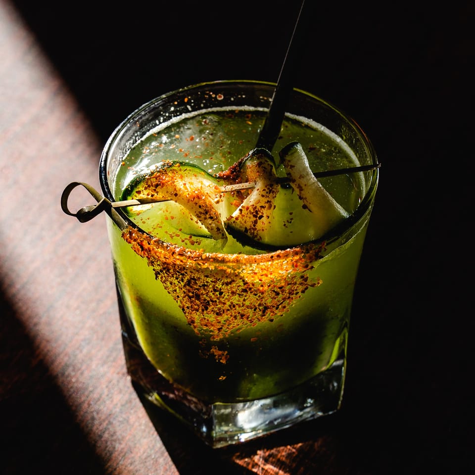 A vibrant green Romesco Mexiterranean Bistro cocktail, garnished with cucumber slices and chili seasoning.