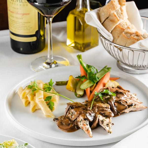 Chicken with mushroom sauce, pasta, and vegetables, with wine and bread in the background.