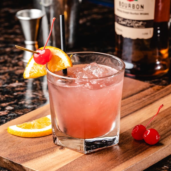 A vibrant pink cocktail with ice, garnished with an orange slice and cherry, on a wooden board.